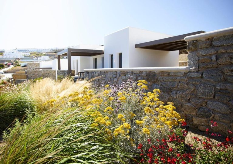 a garden with yellow flowers next to a stone wall at Bonzoe Suites Villas in Mikonos 768x540