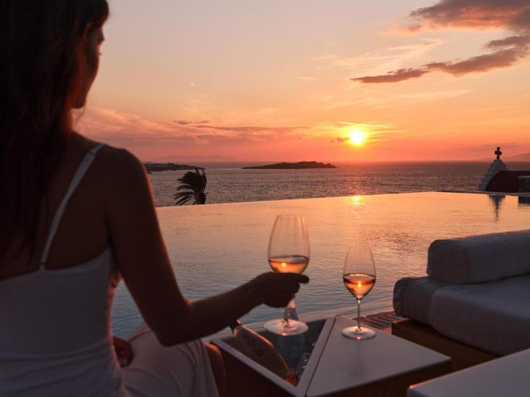 a woman holding two wine glasses in front of a sunset at BillCoo Mykonos The Leading Hotels of the World in Mikonos 768x576